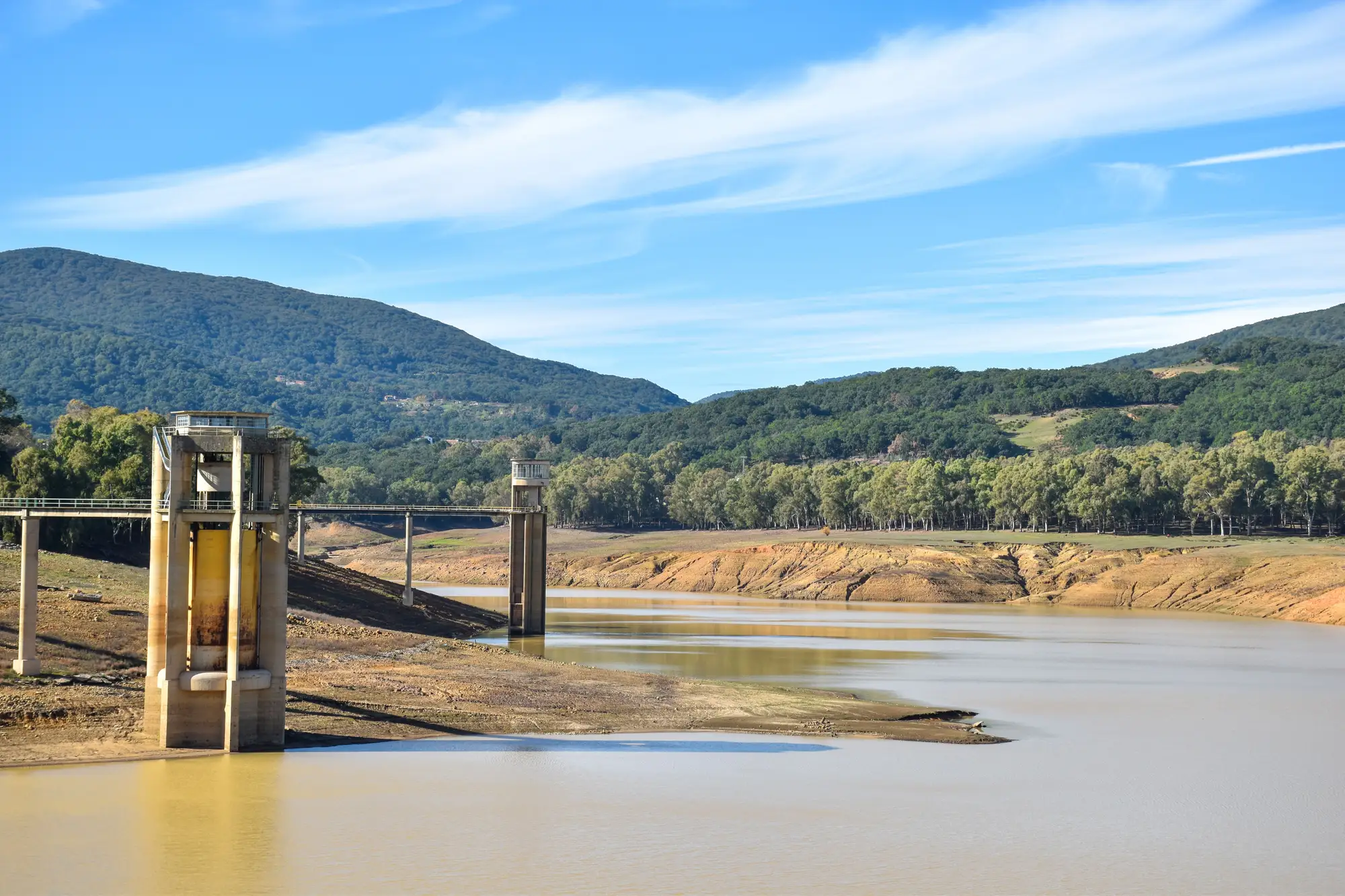 A reservoir late in a dry season, the water pulled back from its intake tower and a bare band of bank showing below the trees