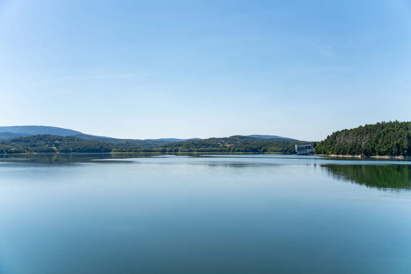 A calm reservoir bordered by forested hills, with trees lining the shoreline and still water reflecting the tree line.